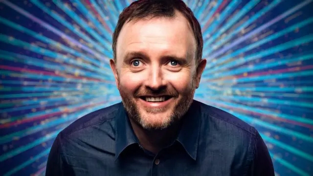 Chris McCausland's Strictly promotional picture. He has short dark hair and a beard and is wearing a blue denim-look shirt