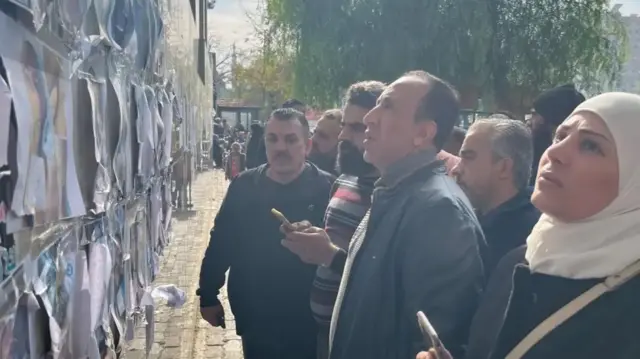 Several men and a woman wearing a white headscard look at a wall covered in photographs of people whose bodies were found at a hospital in Damascus