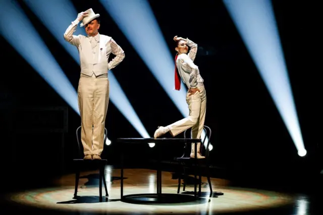 Chris McCausland on the left and Dianne Buswell on the right stands in chairs dressed in white tail coats