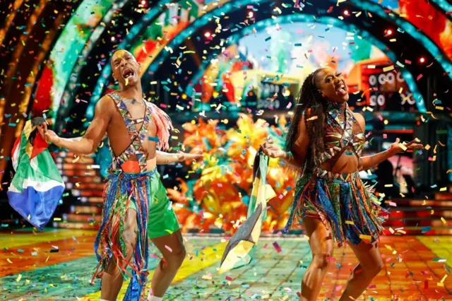 Olympian Montell Douglas and her partner Johannes Radebe performing a carnival-themed dance wearing brightly colours shorts and carrying flags