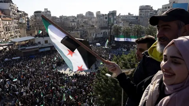 A woman and two men hold an independence flag above a rally in Latakia