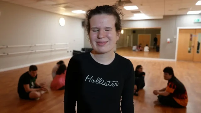 A smiling Amy Griffin wearing a black top stands in a dance studio with a wooden floor and a large mirror