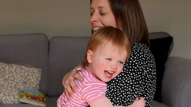 A little red-haired girl in pink striped shirt smiles as she hugs her mum, in polka dot shirt, who smiles back