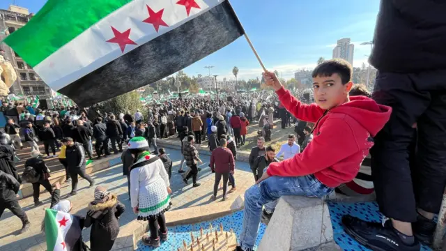 A young child waves an independence flag at a rally in Aleppo. There are large crowds in the background