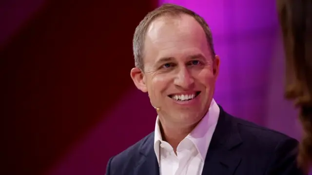 Bret Taylor, show from mid-chest up, wearing dark jacket and white collared shirt, smiling at camera and with a white headset mic on the left side of his face