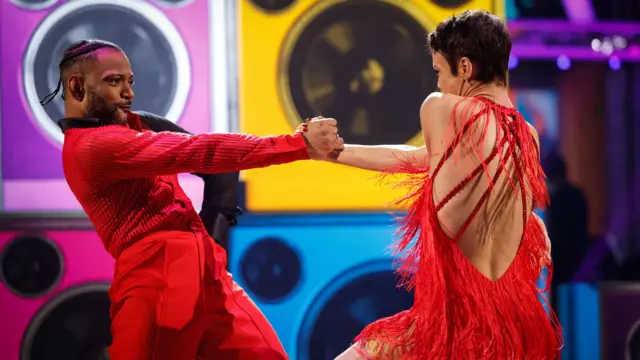 JB Gill is on the left wearing a red shirt and trousers holding the right hand of partner Lauren Oakley who is wearing a red dress