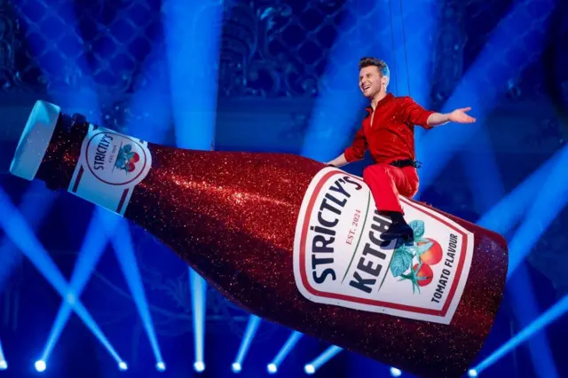 EastEnders actor Jamie Borthwick is sitting on a giant bottle of Strictly's tomato ketchup suspended from the studio ceiling. He is wearing a red shirt and trousers