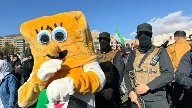 Person wearing Spongebob Squarepants outfit stands and holding flag of the Syrian opposition stands next to two soldiers wearing full black outfits, camouflage body armour and carrying guns