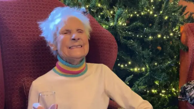 Molly Robinson wears a turtleneck jumper and sits in a red armchair, a Christmas tree is behind her