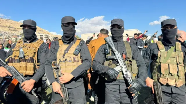 Four people standing in a line wearing full black outfits, camouflage body armour and carrying guns