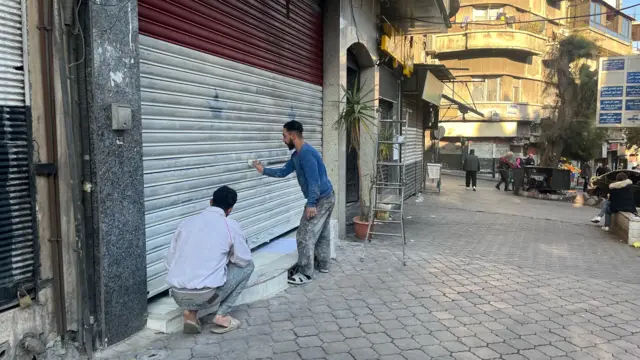 Two men wearing paint-stained clothes paint over red, white and black Syrian flag on shop shutters with white paint