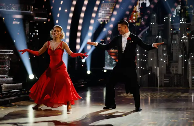 Tasha wearing a red dress with sequins outlining it, while Aljaž mirrors her movement with a black suit