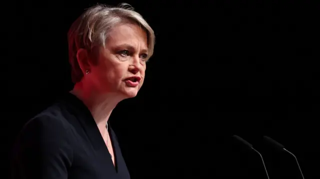 Yvette Cooper speaking into a microphone at a podium on a stage with a black backdrop. She is wearing a black dress and has short grey hair.