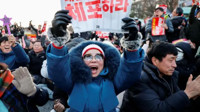 Protestors at a rally to impeach president Yoon. Woman in blue jacket celebrating and holding placard with red Korean characters, surrounded by protestors.