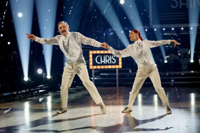 Chris and Dianne holding hands on the dancefloor, wearing all white including white waistcoats, shirts and ties