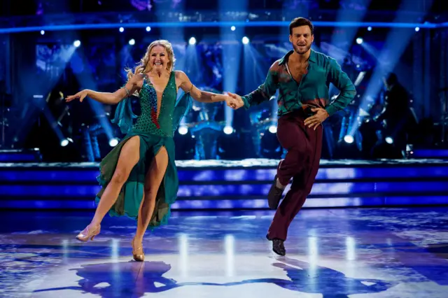 Sarah and Vito jumping in the air, Sarah wearing a sequinned turquoise leotard and flowy skirt, Vito in a turquoise shirt and burgundy straight trousers