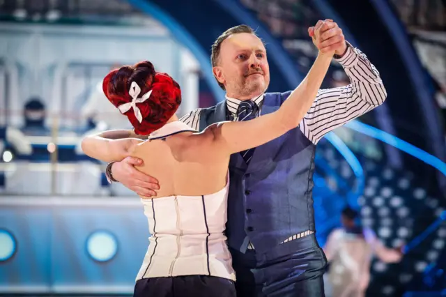 Chris and Di dancing. Their hands are clasped together and they are facing each other. Chris is wearing a white and black striped shirt and a navy blue and white striped tie with a blue waistcoat and trousers.