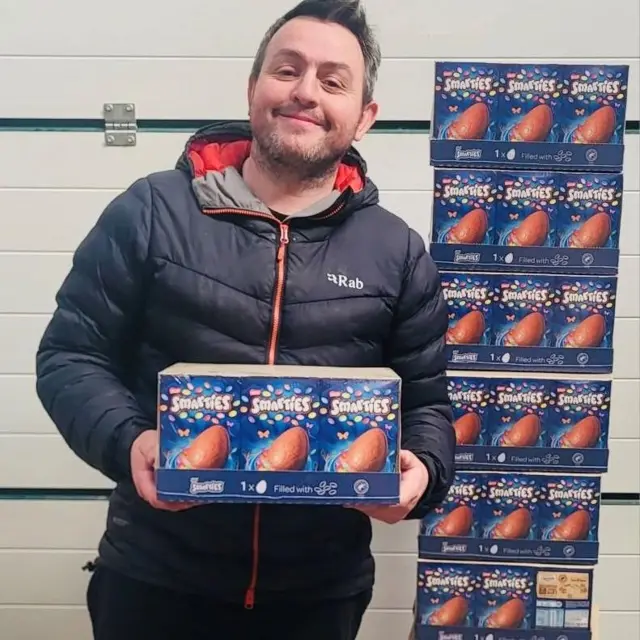 Dan Dafydd holds a crate of Smarties Easter eggs while stood next to a tower of Smarties Easter eggs. Dafydd is wearing a black puffer jacket and smiling at the camera. He has short brown hair.