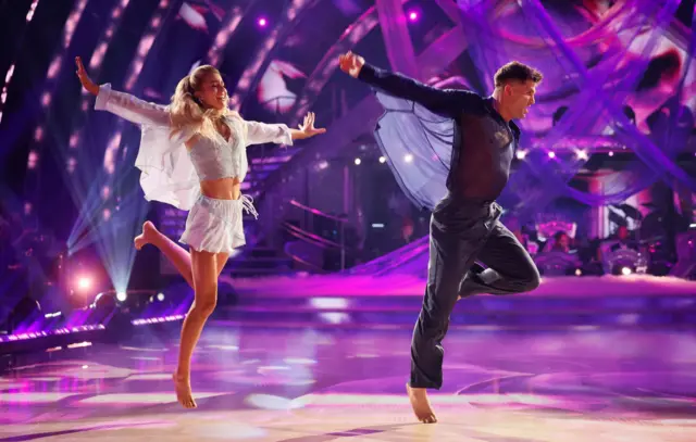 Tasha and Aljaz leaping in the air in mirrored jumps, Tasha in sheer light blue and Aljaz in sheer navy