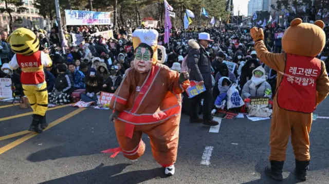 A man wearing a large suit and a mask depicting South Korea's President Yoon Suk Yeol dances in front of a crowd