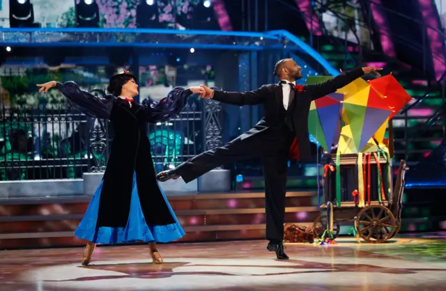 JB Gill and Lauren Oakley, JB on his tip toe with his right leg in the air, with four multicoloured kites on the stage