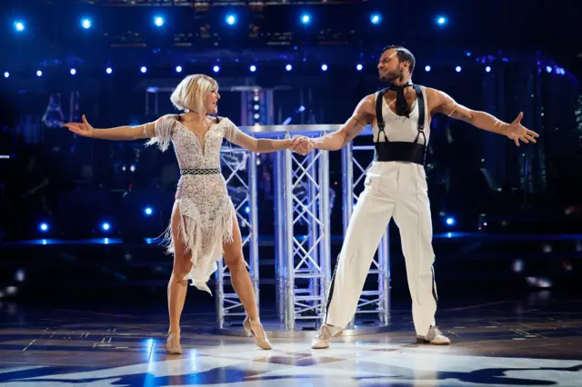 Sarah in a white lace dress with fringe detail, holding hands while looking at partner Vito on the dancefloor