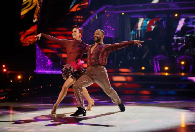 JB lunging on the dancefloor with partner Lauren, both in burgundy outfits, with JB wearing gold sequined trousers and Lauren wearing a big pink rose on her hip