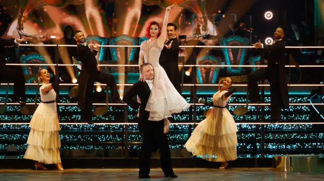 Chris holds up Di while on the ballroom floor. He is wearing a suit and bow tie. Di is wearing a cream-coloured, flowy dress.