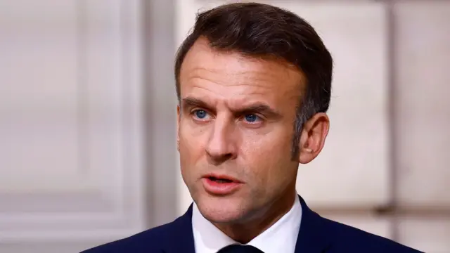 President Emmanuel Macron talking. He is wearing a suit and tie. He has short brown hair and blue eyes.