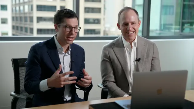 Clay Bavor, left, and Bret Taylor, both sitting at table with a laptop in front of them, doing an interview, with background of high-rise buidlings