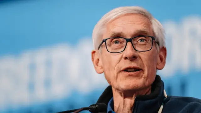 Close-up photo of Wisconsin governor Tony Evers wearing dark blue fleece jacket and blue polo. He stands on stage in front of blurred blue background with unreadable slogan
