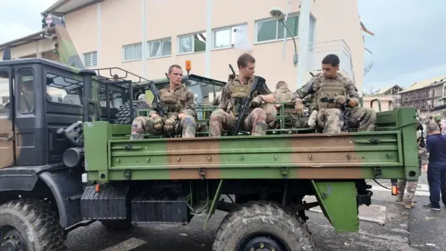 Three uniformed army personnel ride in the tray of a truck through a township