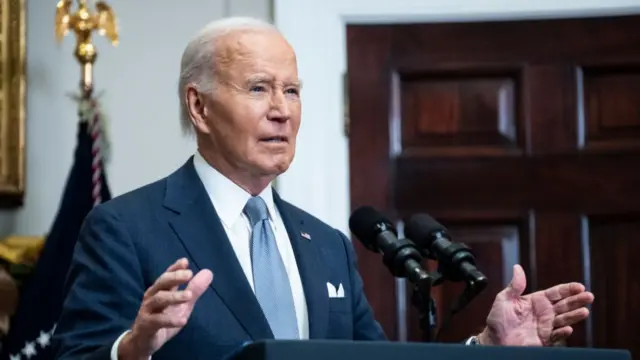 Joe Biden delivers remarks from inside the White House. He stands in front of a lectern, two mics visible on top as he holds his arms open, his hands extending sideways. He's wearing a dark blue suit with a white shirt and light blue tie. A wooden door is visible in the background to his left as is a flag held up by a flag pole topped by a golden eagle