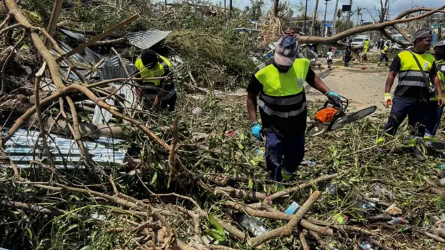 Three recue workers wearing bright colours search through the debris of fallen trees, with one holding a chainsaw