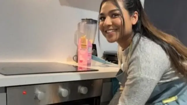 Visha takes a tray of food out of the oven in a kitchen. She has long brown hair and is a wearing a grey jumper and blue apron. There is a pink box of tea and a black blender on the white stone countertop.