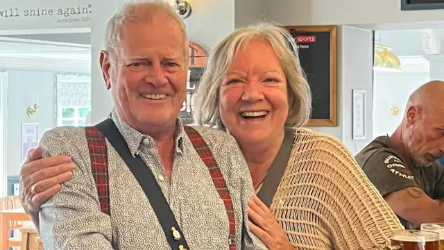 Peter and Leny smiling at a camera, Leny with her arms around Peter, who is wearing a patterned shirt and braces, Leny is wearing a peach crochet top