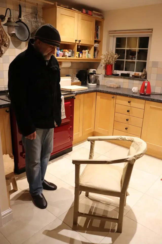 Phil Allen, wearing a coat and hat, looks at a chair he helped make as a child in a kitchen