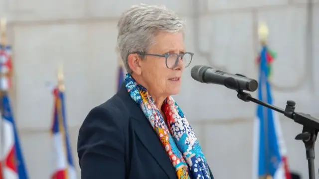 Darrieussecq, a woman with short grey hair, speaks at a lectern, a colourful scarf draped around her neck