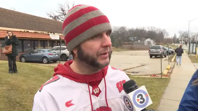 A man in a red hat and a football jersey speaks into a microphone on the street