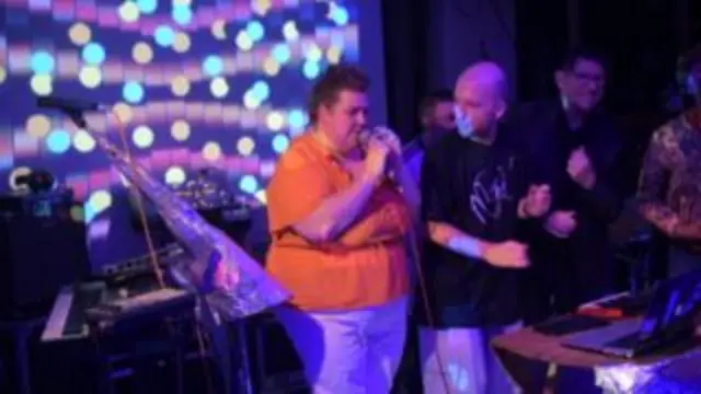 Tina wearing an orange t-shirt and white trousers is singing into the microphone next to Jack, who is dancing, both on a stage.