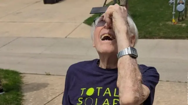 Elderly man in blue t shirt squinting whilst looking at sky through lens. The shirt bears the writing Total solar in lime green. Behind him is a concrete street with visible patches of grass