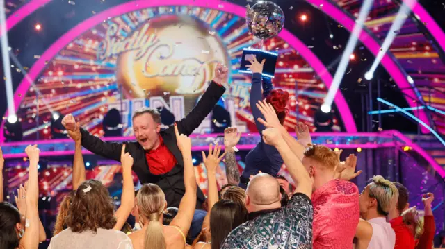 Chris McCausland on someone's shoulders with his arms up in the air, with his partner Dianne holding up the Glitterball trophy