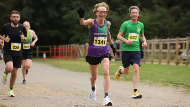 Sarah jumping in the air at the front of four people running, waving to the camera wearing a purple vest and black shorts