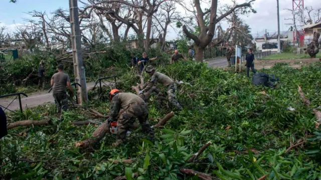 Uniformed army personnel use chainsaws to hack through fallen trees next to a road