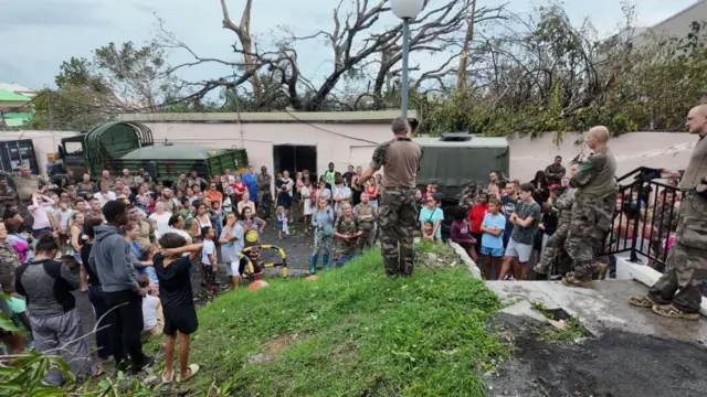 A crowd of around 100 people gather around a uniformed member of the army, who speaks frim a small grassy rise