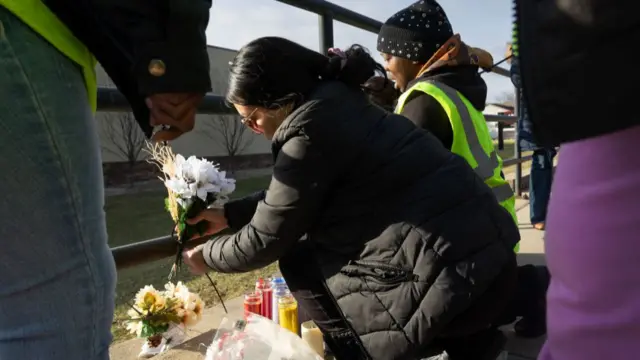 A woman in a black coat attaches a small bouquet of flowers to a metal rail fence. Behind her, another woman in a high vis jacket does the same.