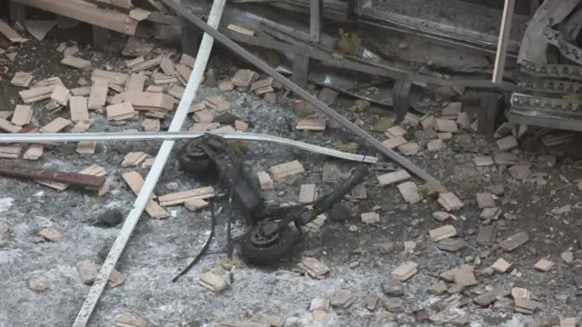 A burned out scooter, apparently scorched black, lies on the floor surrounded by rubble