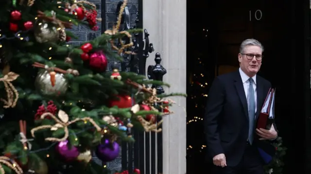 Keir Starmer walks out of Number 10 holding two folders, one red and one black, under his left arm. He's wearing a dark blue suit, patterned blue tie and a white shirt, a Christmas tree partially visible inside the building behind him. To the left of the frame is a close up of another Christmas tree decorated with silver, red and purple baubles