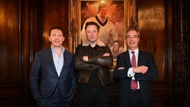 Reform’s new treasury Nick Candy (L) in dark blue suit and unbuttoned blue shirt with his hands in his pockets, X owner Elon Musk (C) in brown leather jacket, black shirt and trousers with his arms crossed in front of his chest; Nigel Farage (R) in black suit, white shirt and red patterned tie smiling, his arms crossed. Hanging behind them is a portrait of a young Donald Trump in a white jumper, shirt and trousers on a wooden-panelled wall