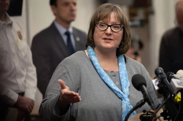 Barbara Wiers speaks at a press conference. She has short, light brown hair, and wears glasses.